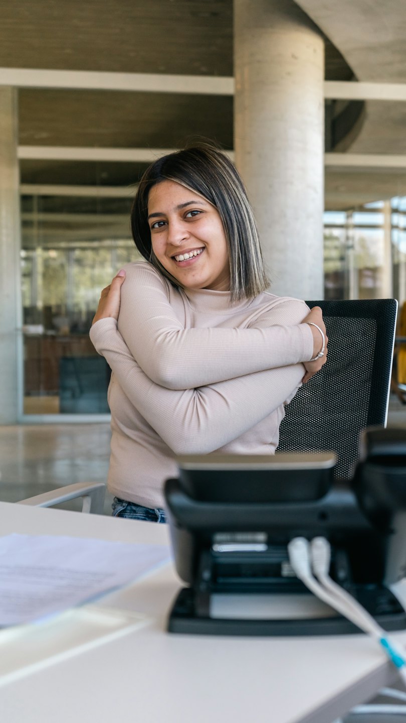 Woman in a beige top sits smiling at a desk.